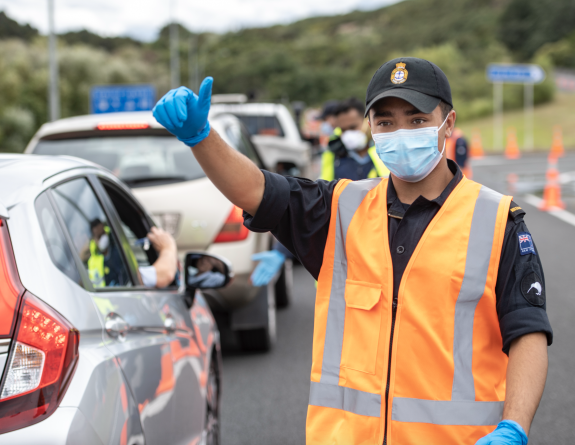 A sailor in high-vis vest and face mask waves vehicles through a COVID checkpoint.