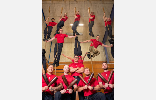 Physical Training Instructors at the Fleet Gym. Some of the PTIs hold brown clubs in the foreground while others are suspended from ropes in varied positions in the background, showcasing their strength and agility.