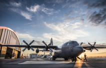 C-130H(NZ) Hercules stationary on flightline at RNZAF Base Auckland. With hangars in the background, a large puddle mirrors the aircraft and blue sky above.