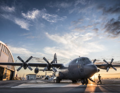 C-130H(NZ) Hercules stationary on flightline at RNZAF Base Auckland. With hangars in the background, a large puddle mirrors the aircraft and blue sky above.