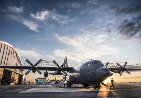 C-130H(NZ) Hercules stationary on flightline at RNZAF Base Auckland. With hangars in the background, a large puddle mirrors the aircraft and blue sky above.