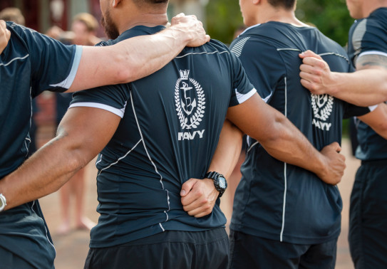 Royal New Zealand Navy sailors stand side-by-side, with arms on each others backs. The sailors are wearing t-shirts with the Navy logo on the back of them.